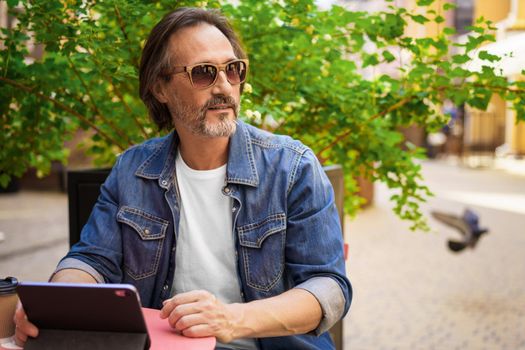 Middle aged man looking away sitting at cafe or street restaurant while working outdoors using digital tablet. Handsome mature freelancer man enjoying coffee outdoors while having group chat.