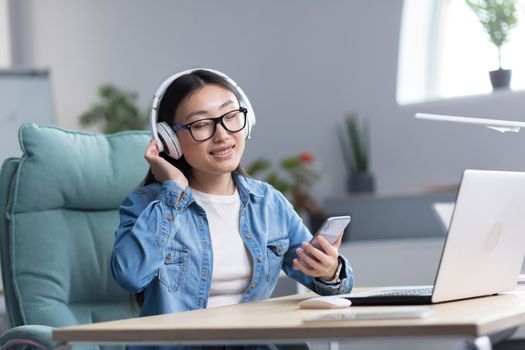 Young beautiful Asian woman listening to music in office, using phone and music app, businesswoman with headphones resting.