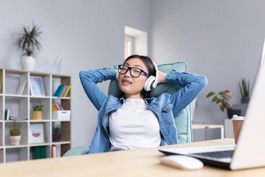 Young beautiful office worker relaxing sitting at the desk, Asian woman listening to music in headphones during work break.