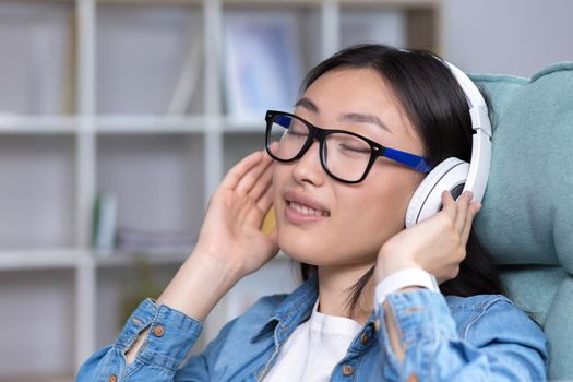 Close-up portrait of a young Asian woman with closed eyes, listening to music and relaxing in with white headphones.