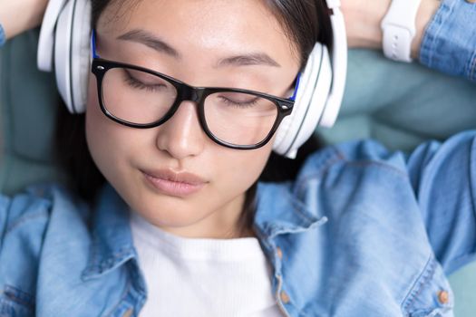 Close-up portrait of a young Asian woman with closed eyes, listening to music and relaxing in with white headphones,