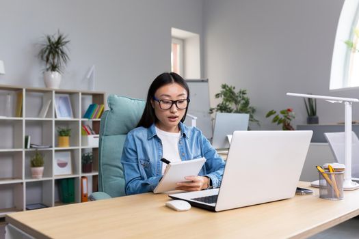 Young Asian woman studying online while sitting in office, writing lecture notes in notebook, using laptop for studying.