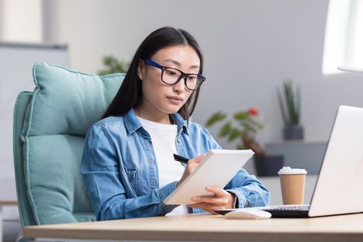 Young Asian woman studying online while sitting in office, writing lecture notes in notebook, using laptop for studying.