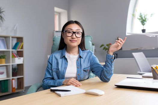 Young beautiful Asian woman talking on video call, business woman working in modern office, smiling and looking at web camera, online conference.