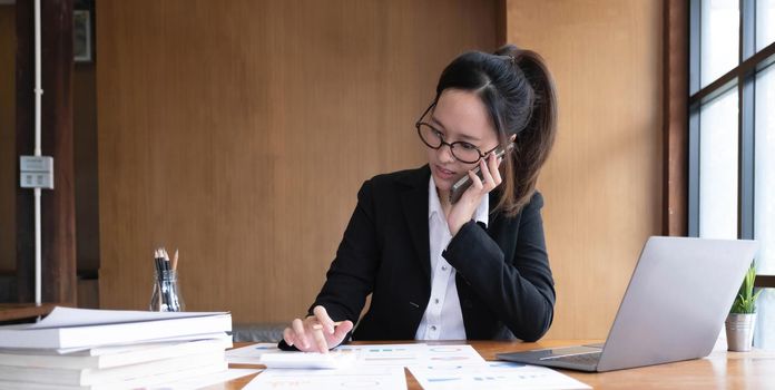 Happy young Asian business woman sitting smiling and talking on mobile phone in office..