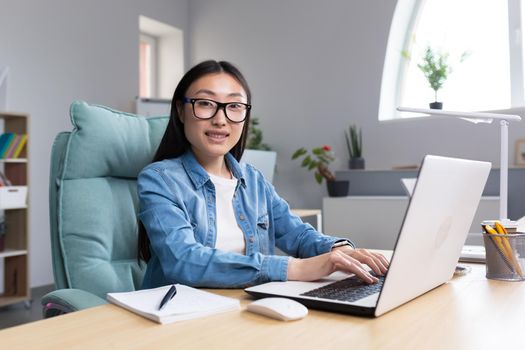 Young business woman working in office at laptop, asian smiling looking at camera, successful and happy employee.