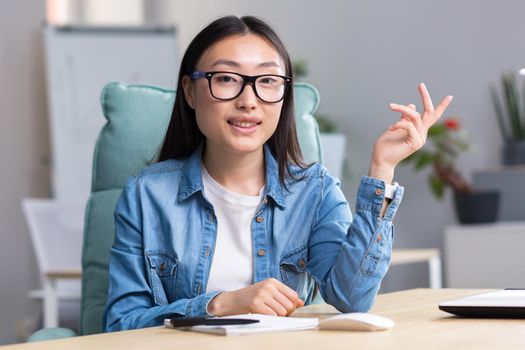 Young beautiful Asian woman talking on video call, business woman working in modern office, smiling and looking at web camera, online conference.