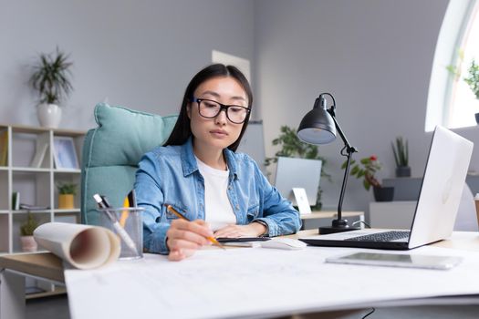 Asian designer working in office, with blueprints, woman at work in casual clothes.