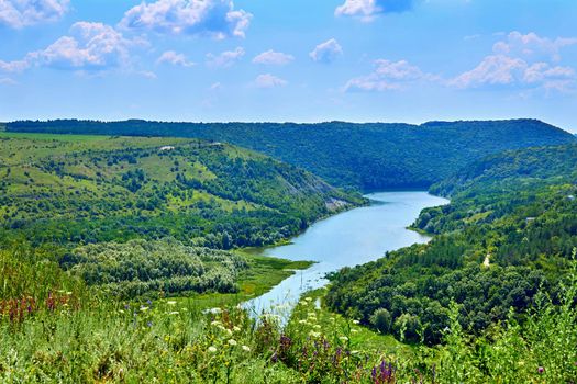 a large natural stream of water flowing in a channel to the sea, a lake, or another such stream.Summer river flowing among lush flowering green hills. Tovtry National Park Ukraine