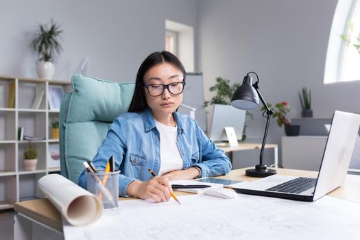 Asian designer working in office, with blueprints, woman at work in casual clothes.
