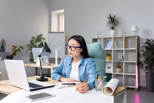 Asian designer working in office, with blueprints, woman at work in casual clothes.