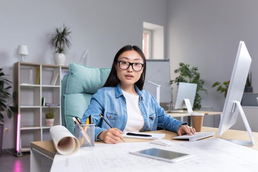 Portrait of young designer woman in modern office at work, successful Asian woman in glasses looking at camera.