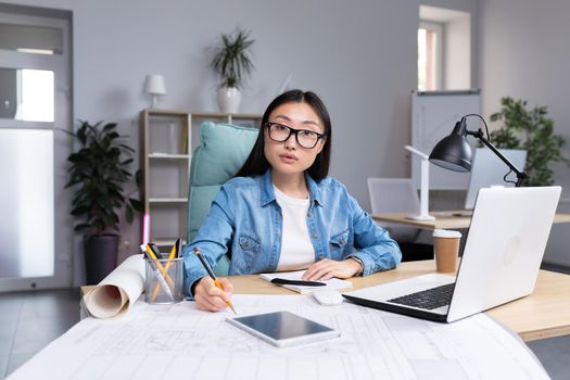 Portrait of young designer woman in modern office at work, successful Asian woman in glasses looking at camera.