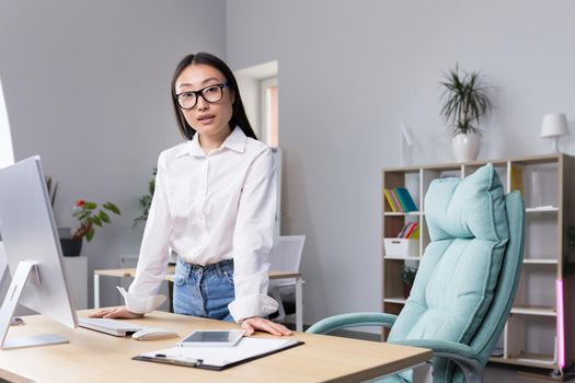Successful Asian business woman at workplace in office looking at camera, portrait of strong leader.