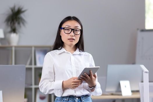 Portrait of a young Asian businesswoman standing in the office and looking at the camera, secretary wearing a white shirt and glasses, employee holding a tablet.