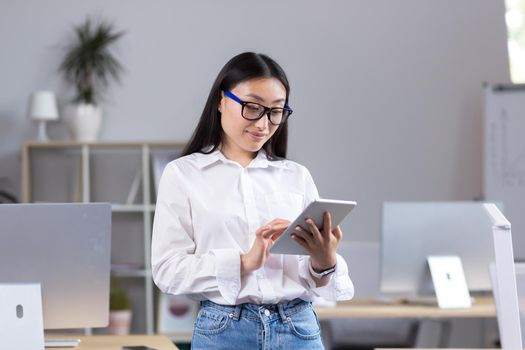 Young beautiful Asian teacher working with tablet, in classroom with computers at school, woman in white shirt and glasses reads from tablet