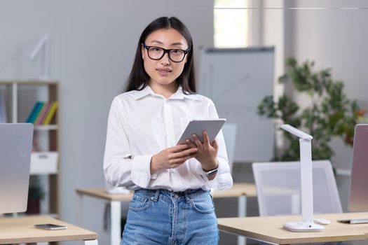 Portrait of a young Asian businesswoman standing in the office and looking at the camera, secretary wearing a white shirt and glasses, employee holding a tablet.