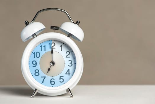 Seven o'clock on the alarm. A white alarm clock is on a white table. The clock hand points to 7 o'clock. Time to change to summer or winter time. Set an alarm for 7:00 or 19:00. copy space