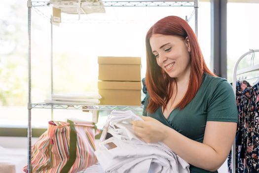 young woman entrepreneur, restocking trousers on the shelves of her clothing shop. woman owner of a small business taking inventory. work and business concept.natural light from window, background with clothes racks and clothes.