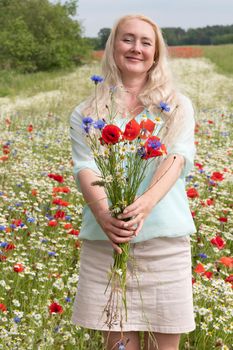 a beautiful middle-aged blonde woman stands among a flowering field of poppy, daisies, Cornflowers, and holds a bouquet of wild flowers and laughs. High quality photo