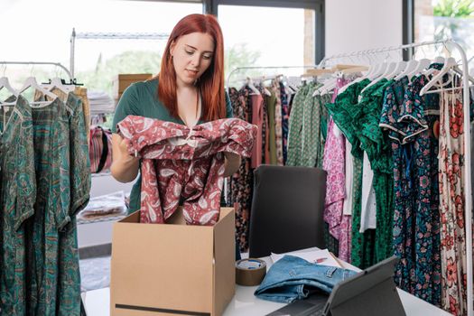 young woman entrepreneur smiling, packing clothes in a box for a shipment of her online clothing shop. woman owner of a small online business. work and business concept. natural light from window, background with clothes racks and clothes.