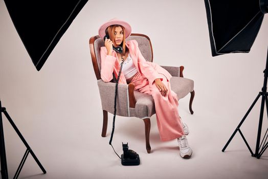 Stylish young mixed race female posing in trendy pink clothes sitting on a chair and making a call with a retro telephone for a studio photoshoot. Female model on the set of an editorial fashion shoot.