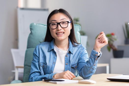 Young beautiful Asian woman talking on video call, business woman working in modern office, smiling and looking at web camera, online conference.