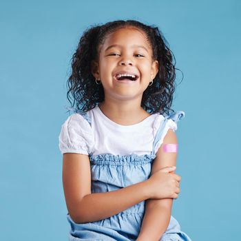 Happy vaccinated little girl kid showing arm with adhesive bandage after vaccine injection standing against a blue studio background. Advertising vaccination against coronavirus. Child immunisation.