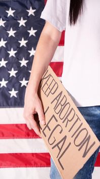 Young woman protester holds cardboard with Keep Abortion Legal sign against USA flag on background. Girl protesting against anti-abortion laws. Feminist power. Equal opportunity Womens rights reedom