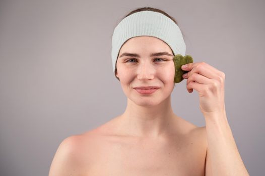 Portrait of a young woman massages her face with a gouache scraper on a white background