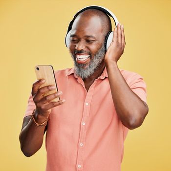 One mature african american man listening to music using wireless headphones while isolated against a yellow background. Happy man with a grey beard smiling while streaming on his phone in studio.