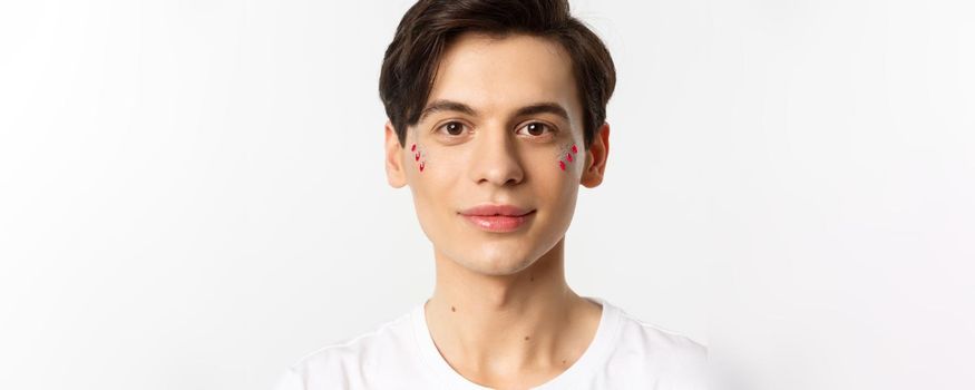 People, lgbtq and beauty concept. Close-up of happy queer guy with applied lip gloss and glitter, smiling and looking at camera, standing over white background.