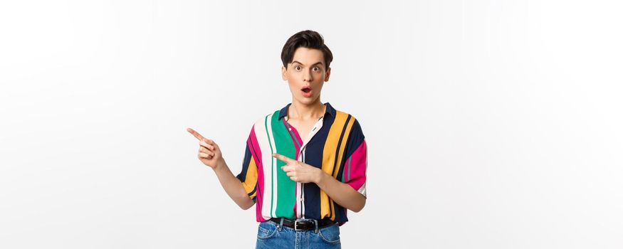 Amazed young queer man pointing fingers left, staring at camera with disbelief, asking question about product, standing over white background.