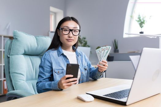 Portrait of young beautiful freelancer woman looking at camera and smiling holding phone and american money dollars, online win.