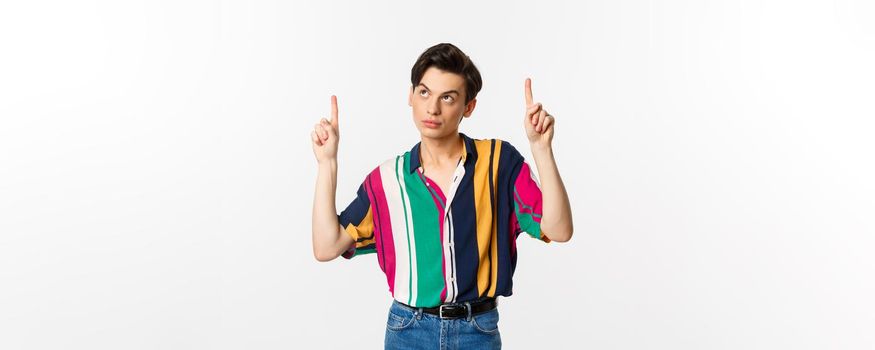 Attractive gay man looking thoughtful, pointing fingers up at logo, standing over white background. Copy space