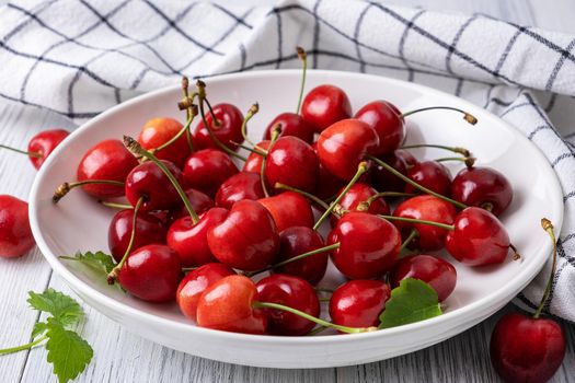 Ripe juicy sweet cherry lies on a white porcelain plate on a light wooden surface. Summer healthy food. Selective focus.