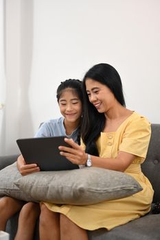 Asian mom and daughter having fun browsing internet on digital tablet together on sofa.