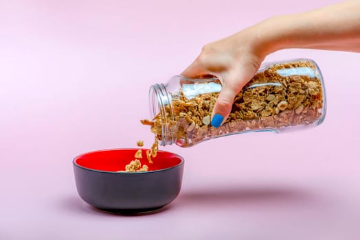 Muesli on a pink background. Pour dry muesli flakes by hand into a bowl. Pouring muesli from a glass jar into a black bowl when making cereals with muesli