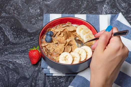 Muesli. Breakfast, healthy food and diet. Muesli with milk and fruit in a plate on a black marble top. Woman's hand with a spoon.
