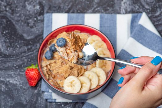 Muesli. Breakfast, healthy food and diet. Muesli with milk and fruit in a plate on a black marble top. Woman's hand with a spoon.