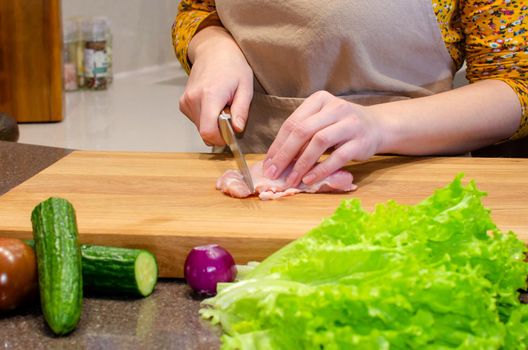 Woman in an apron in the kitchen slicing chicken fillet on an oak board. Vegetables in the kitchen, marble countertop