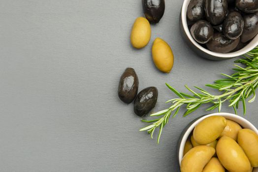 Black and green olives. Bella di cerignola Italian olives. Colored olives and a sprig of rosemary lie on a black stone countertop. Culinary banner or poster for advertising with place for text.