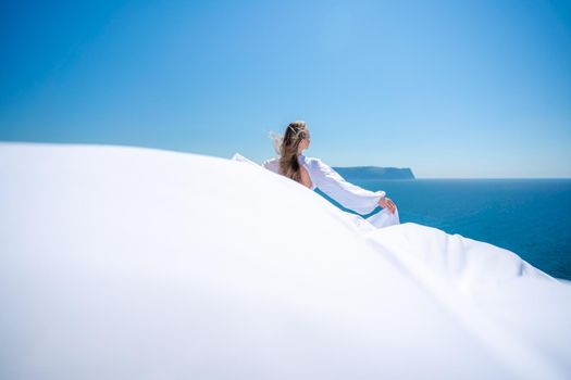Blonde with long hair on a sunny seashore in a white flowing dress, rear view, silk fabric waving in the wind. Against the backdrop of the blue sky and mountains on the seashore