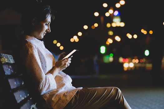 Young woman with smartphone sit on the bench at night, blurred lights of building background on the street