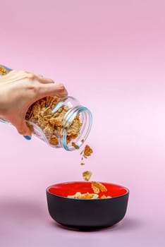 Muesli on a pink background. Pour dry muesli flakes by hand into a bowl. Pouring muesli from a glass jar into a black bowl when making cereals with muesli