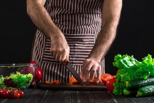 Young home cook man in apron slicing carrot with kitchen knife. Men's hands cut the carrot to make a salad on black background. Healthy food concept