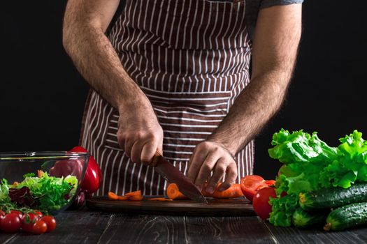 Young home cook man in apron slicing carrot with kitchen knife. Men's hands cut the carrot to make a salad on black background. Healthy food concept