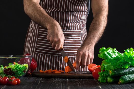 Young home cook man in apron slicing carrot with kitchen knife. Men's hands cut the carrot to make a salad on black background. Healthy food concept