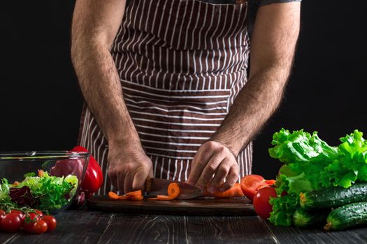 Young home cook man in apron slicing carrot with kitchen knife. Men's hands cut the carrot to make a salad on black background. Healthy food concept