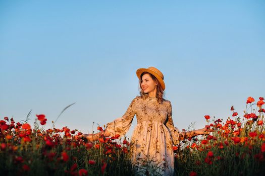 a girl in a dress, in a hat walks in a field with poppies at sunset.